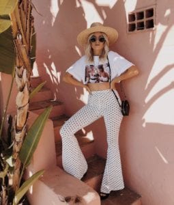 Tessa (@tessalindsaygarcia) is a fashion blogger from Vancouver that frequents the Okanagan; she is wearing a white crop top with high-waisted flare pants and shades and a hat in the shade of palm leaves